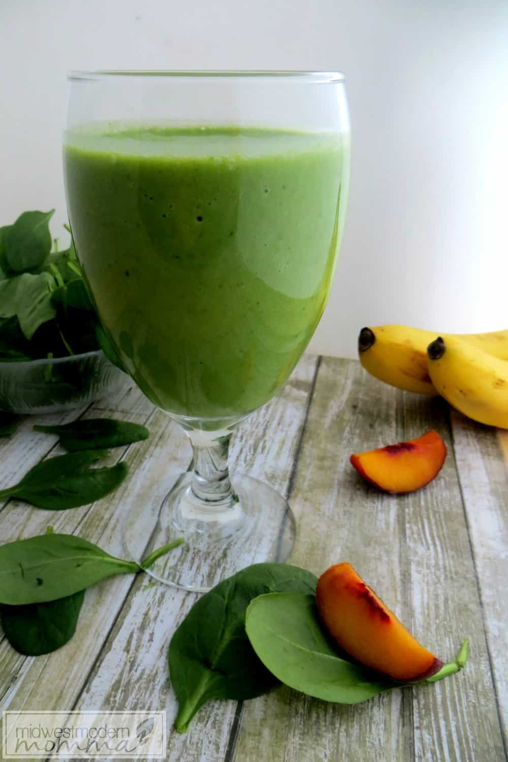 Green smoothie in a glass cup on a wooden surface with spinach, peach slices, and 2 bananas in the background.