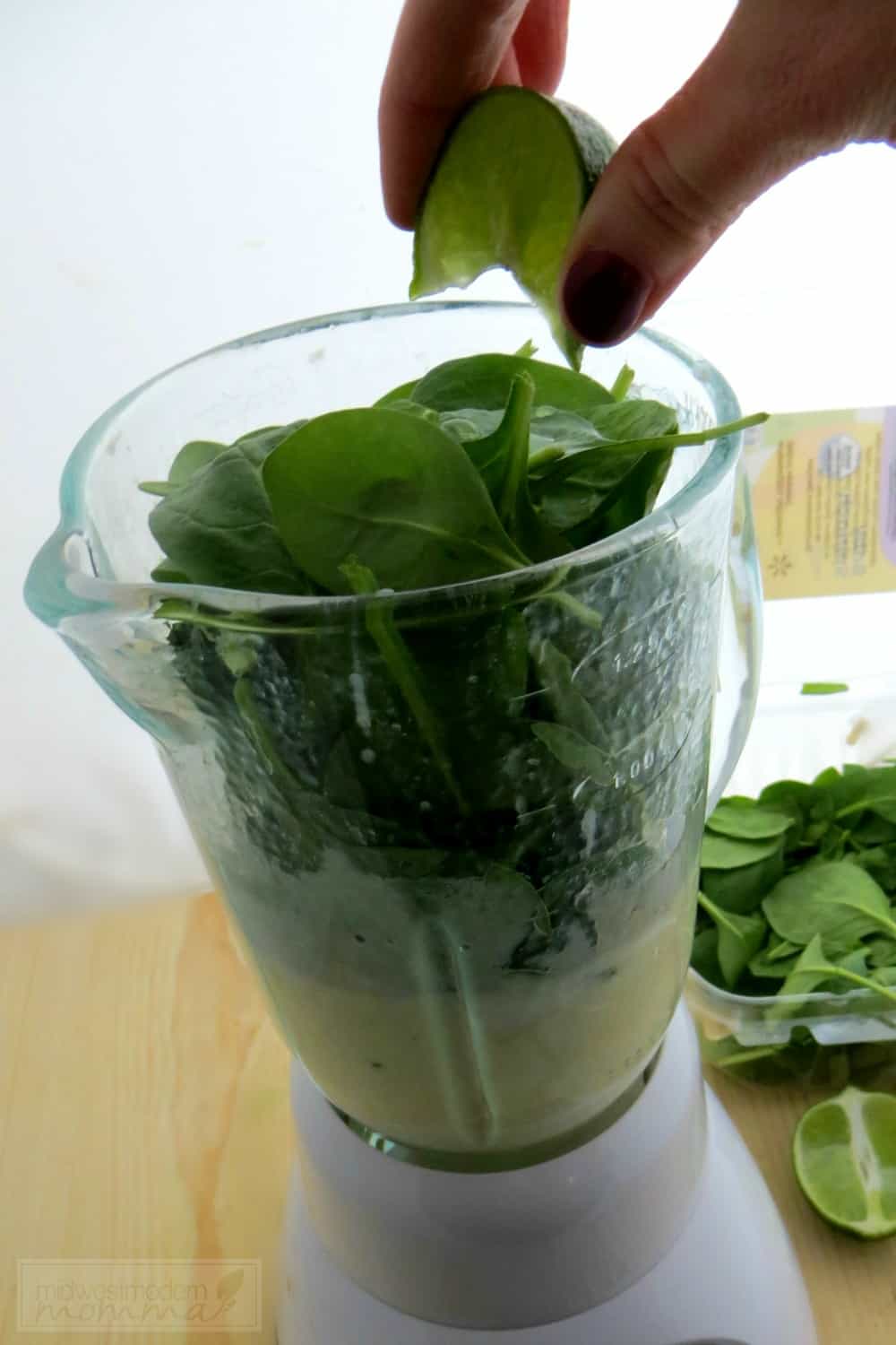 Hand squeezing a lime slice into a blender pitcher full of spinach leaves and a light coloured liquid.