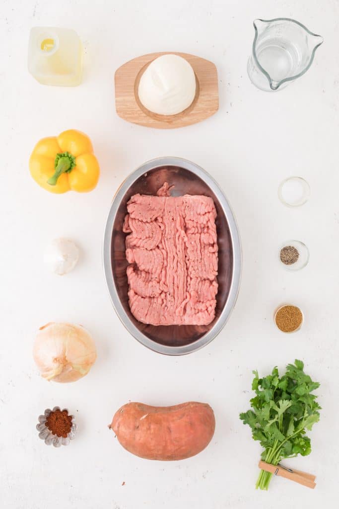 Ground turkey in a metal plan surrounded by ingredients for a skillet dinner, including yellow bell pepper, onion, garlic, cilantro, a sweet potato,  a measuring cup, a ball of fresh mozzarella, and various spices in little bowls.