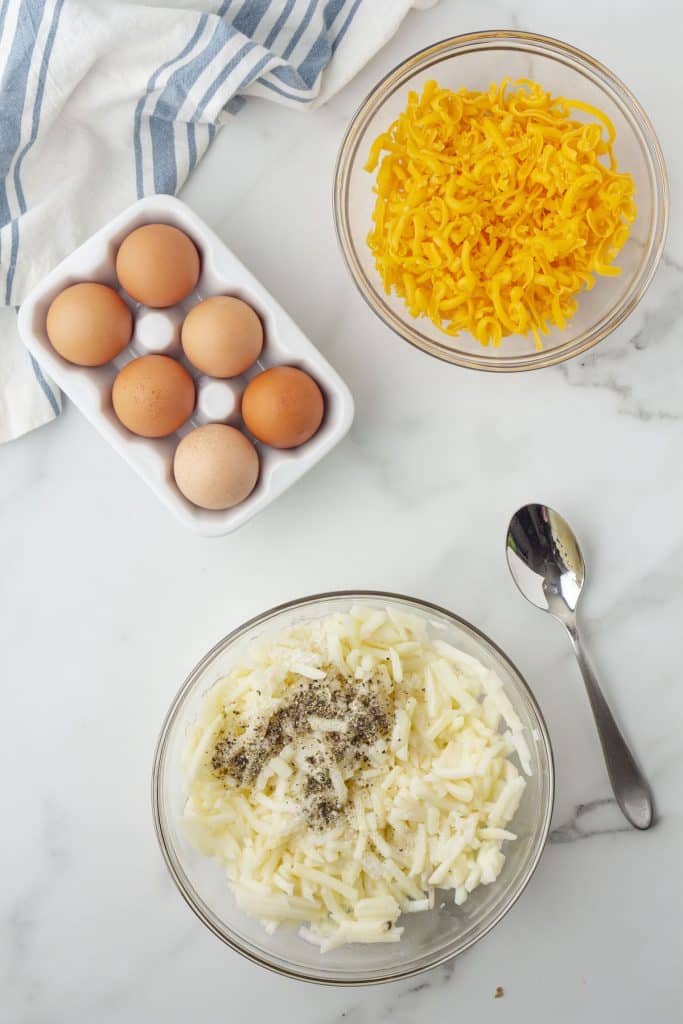 A bowl of seasoned hash browns below a container of eggs and a bowl of shredded cheese.
