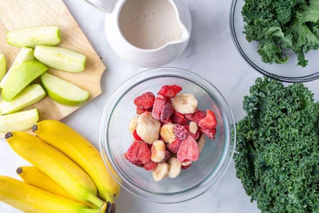 Cut apples, bananas, strawberries, kale, and a pitcher of almond milk on a counter.
