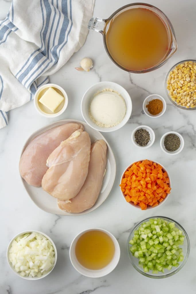 Chicken breasts, celery, carrots, corn, and other ingredients for chicken and dumplings on a marble countertop.