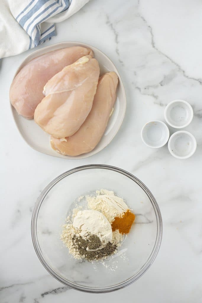 A glass bowl of spices next to three empty pinch pots and a plate of raw chicken breasts.