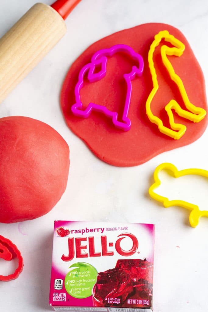 A ball of red jello playdough with cookie cutters, a rolling pin, and a box of raspberry jello.
