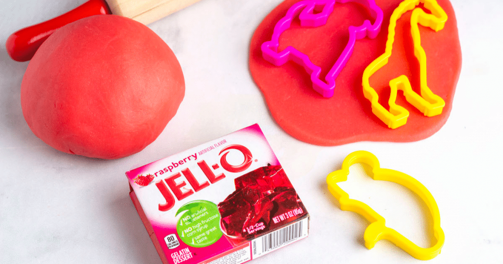 A ball of red jello playdough with cookie cutters and a box of raspberry jello.
