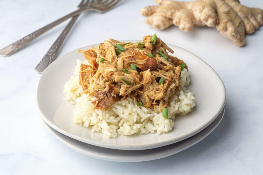 White plate with chicken teriyaki on a bed of rice, with two forks and ginger root in the background.