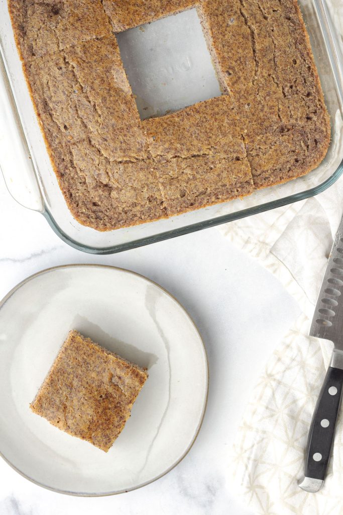 A glass pan of paleo cornbread, cut into large squares with a single square on a white plate with a knife to the side on a light coloured kitchen towel.