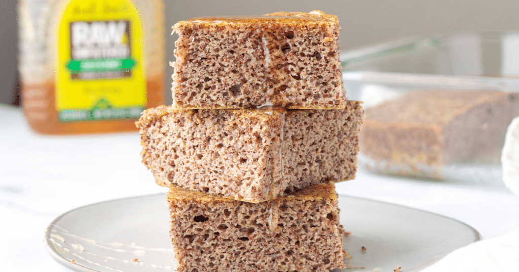 3 squares of paleo cornbread stacked on a light coloured plate with honey dripping off the sides of the cornbread onto the plate., a bottle of honey and a pan of more cornbread in the background.