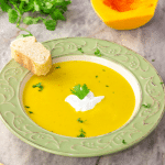 A bowl of savory butternut squash soup topped with shredded with fresh cilantro and yogurt with a cut squash in the background.
