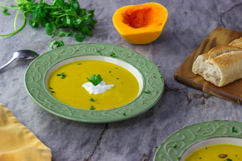 A bowl of savory butternut squash soup topped with shredded with fresh cilantro and yogurt with a cut squash in the background.