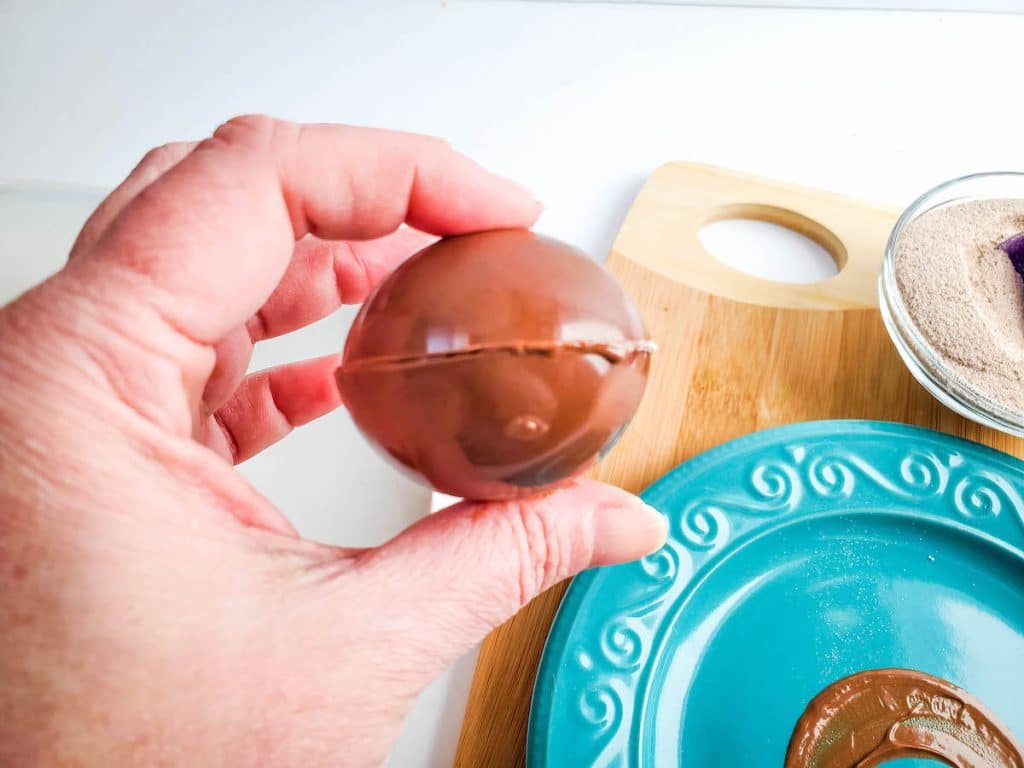 A hand holds 2 halves of chocolate shell together with a blue plate in the background.
