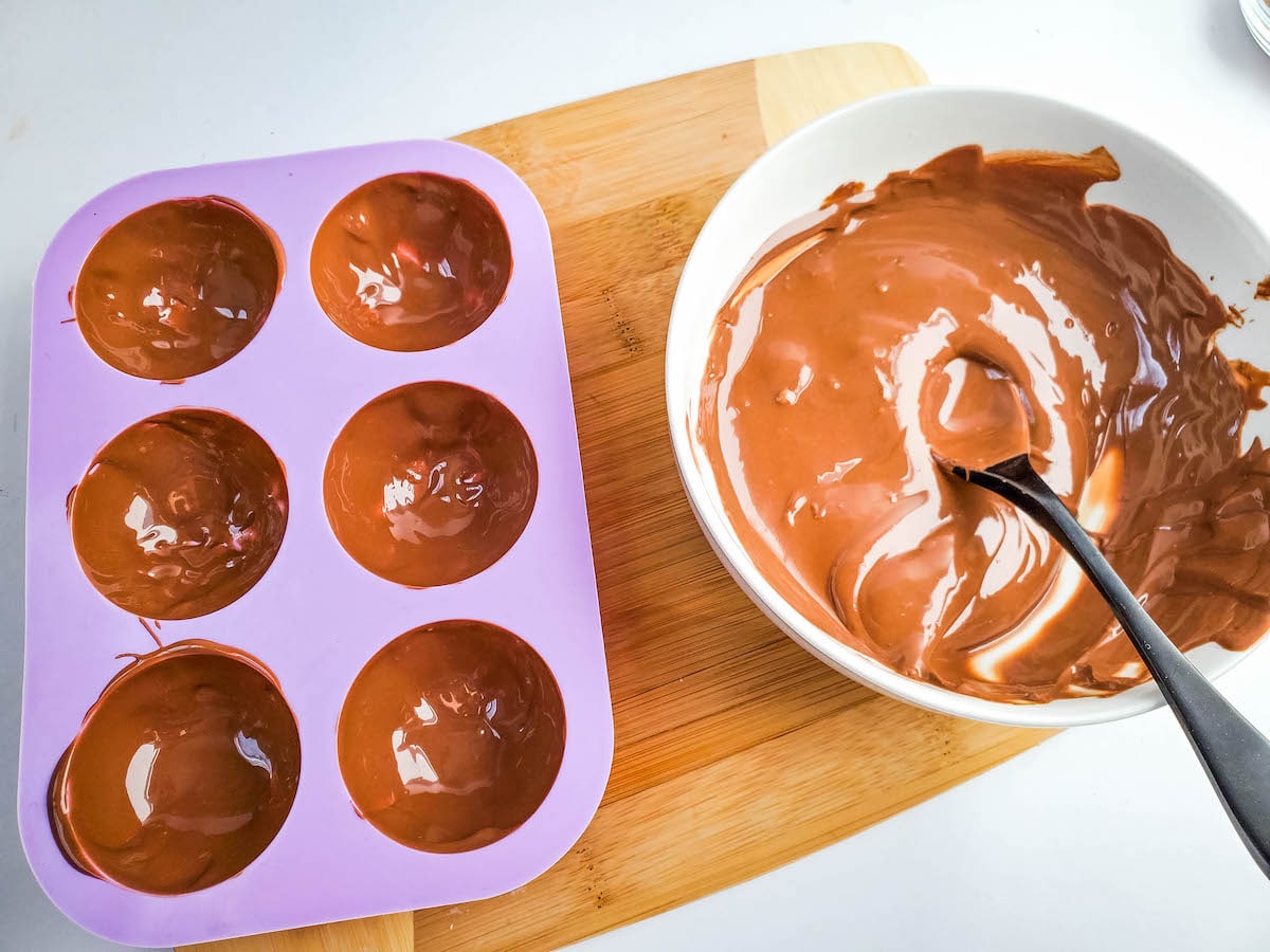 A half-full bowl of melted chocolate with a spoon next to a purple silicone mold with melted chocolate in each cavity, on a wooden cutting board.