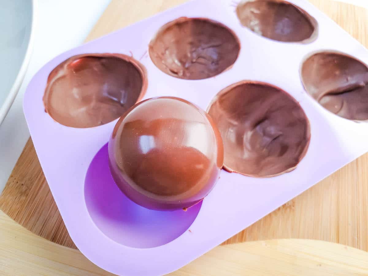 A purple silicone mold with 6 chocolate shells on a wooden cutting board.