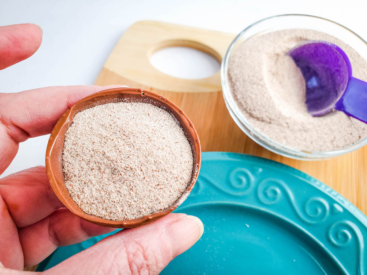 A hand holding a chocolate shell with cappuccino powder, with a blue plate and glass bowl with more cappuccino powder and a purple scoop in the background.