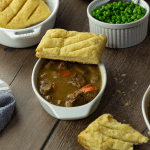 A bowl of steak pie on a wooden table with a side dish of peas.