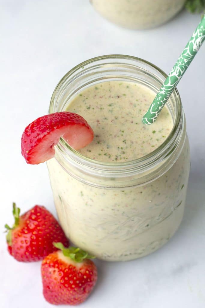 Mason jar full of a light coloured smoothie with green flecks, with a green straw sticking up and a sliced strawberry on the rim.