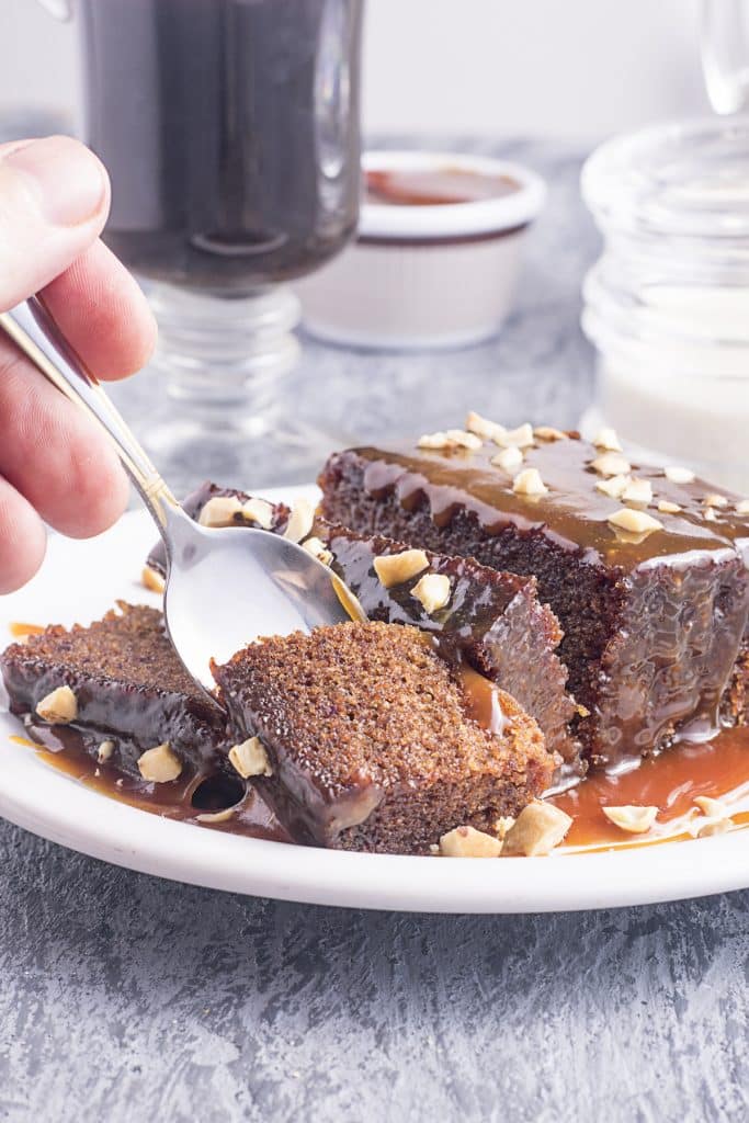 Close-up of a white plate on a gray distressed table, showcasing slices of sticky toffee pudding drenched in caramel sauce. A hand is seen holding a spoon, taking a bite from the cake. In the slightly blurred background, there's a clear glass mug filled with a dark liquid, a white ramekin with extra toffee sauce, and a clear container holding cream.