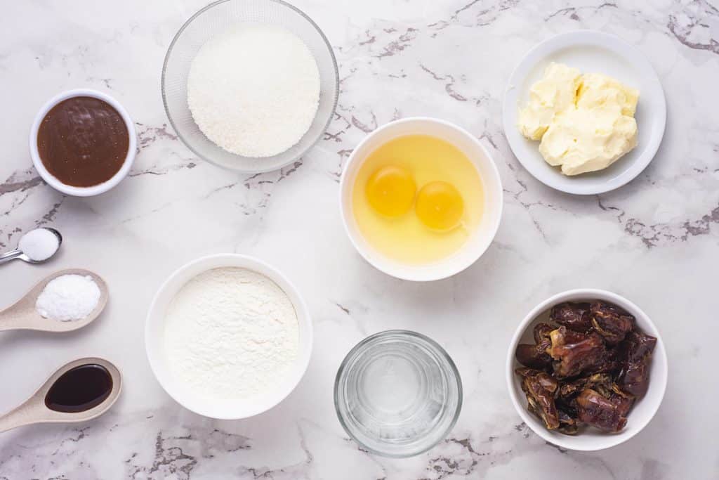Flatlay photograph of ingredients for making dessert. Features toffee sauce in a white dish, separate bowls of flour and sugar, two eggs in a bowl, softened butter on a saucer, pitted dates in a white bowl, a glass of water, and measuring spoons holding a dark liquid and two distinct white powders.
