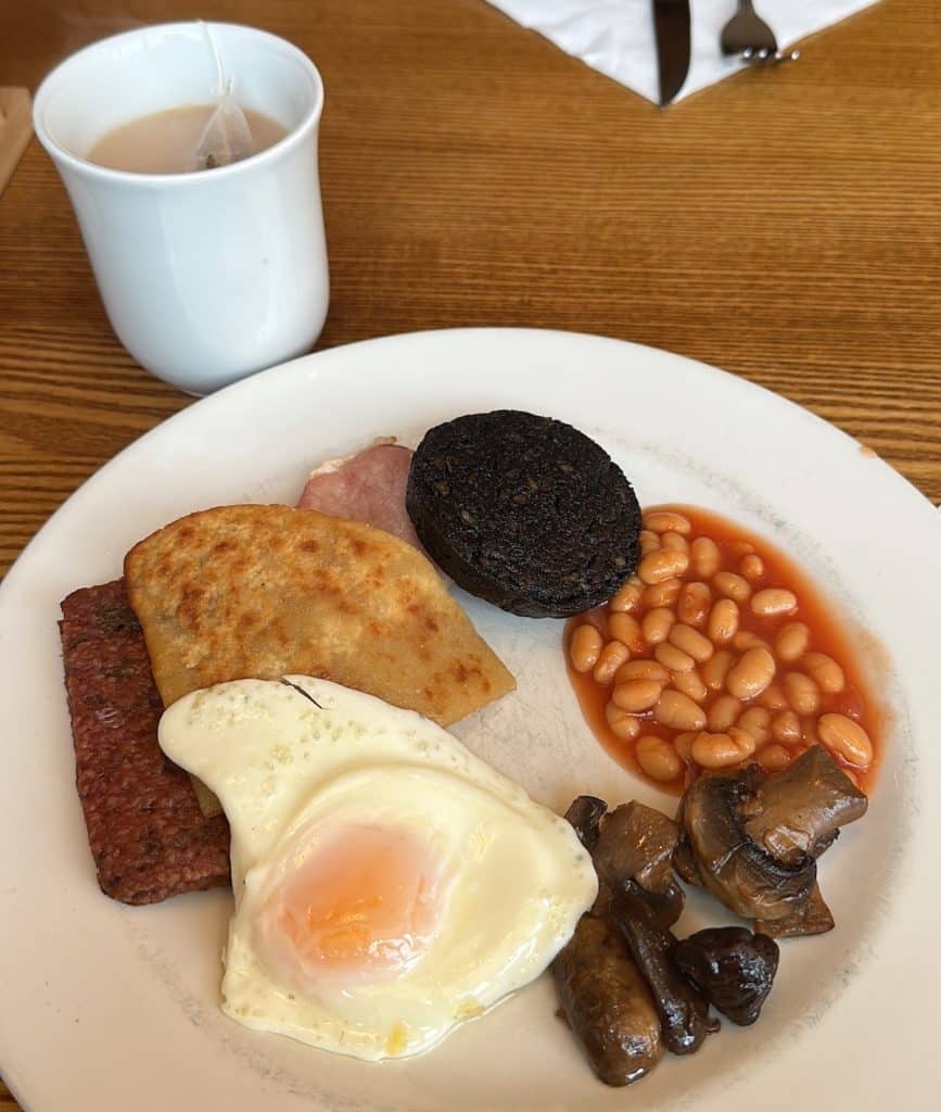 A cup of British tea with milk on a wooden table, next to a white plate with square sausage, tattie scone, a rasher of bacon, slice of black pudding, baked beans, fried mushrooms, and a fried egg.