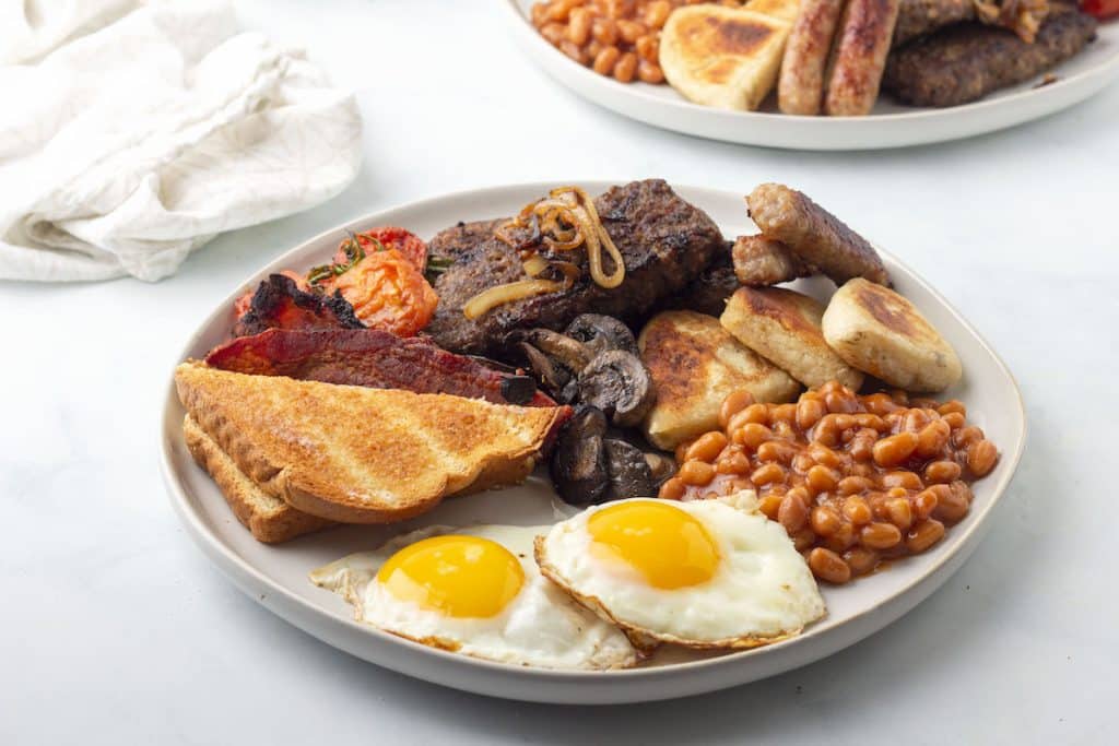 A plate of breakfast food with eggs, beans, lorne sausage, tattie scones, tomatoes, mushrooms, bacon, pork sausage links, and toast.