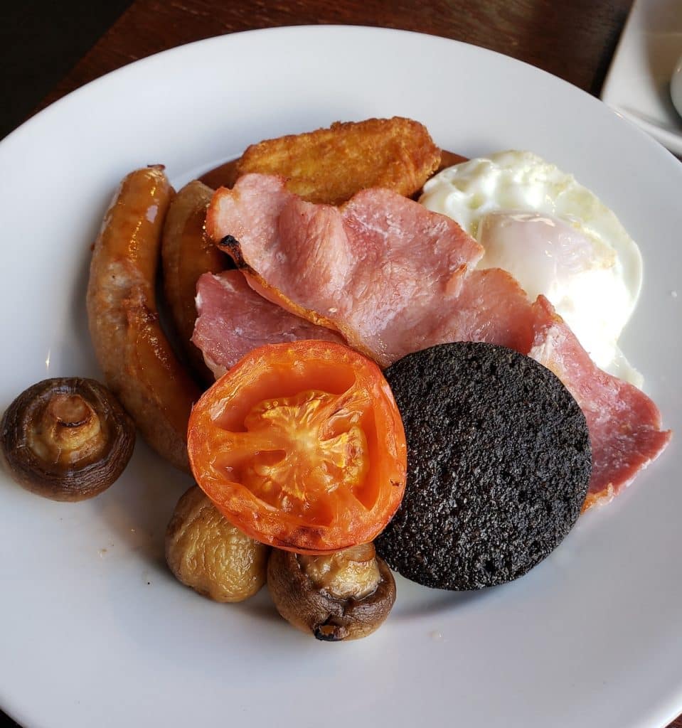 A white plate with fried mushrooms, fried tomatoes, a slice of black pudding, 2 rashers of bacon, 2 sausage links, a fried egg, and a hashbrown.