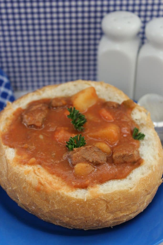 Beef stew in a bread bowl on a blue plate with salt & pepper shakers in the background.