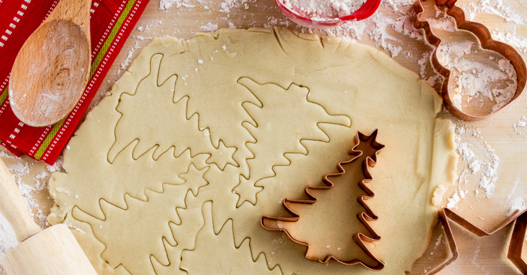 Cookie dough rolled out on a wooden surface, with Christmas trees cut into the dough and various cookie cutters sitting nearby.