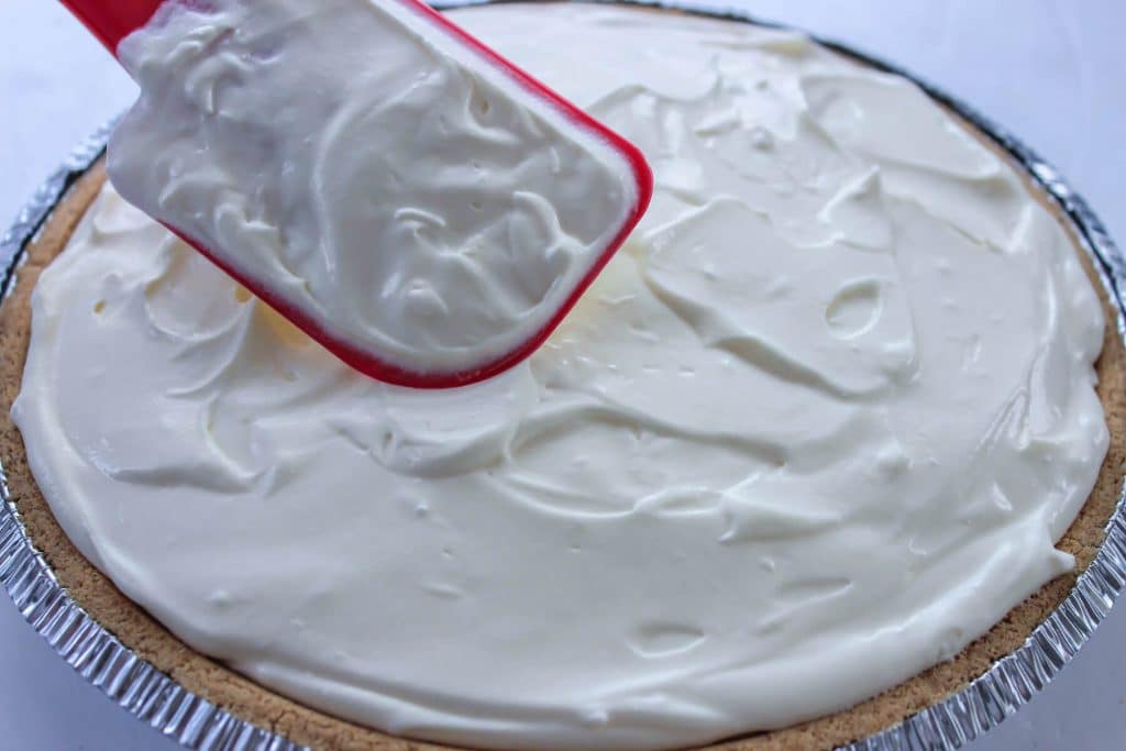 A red spatula smoothing the top of a banana cream pie.