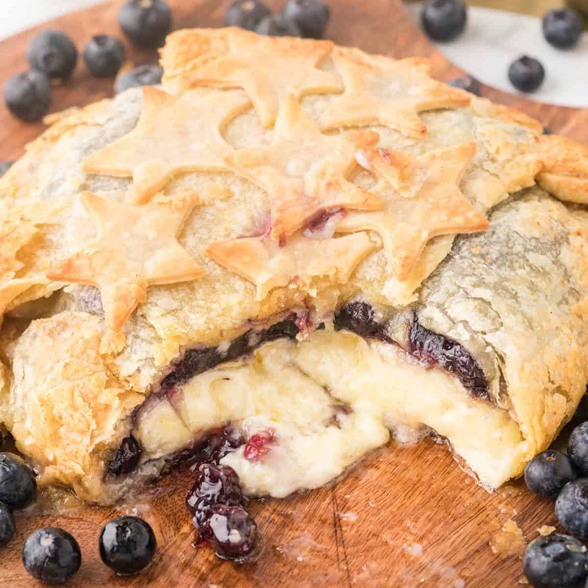 Melty brie topped with blueberry pie filling, oozing out of a puff pastry crust onto a wooden board, with blueberries in the background.