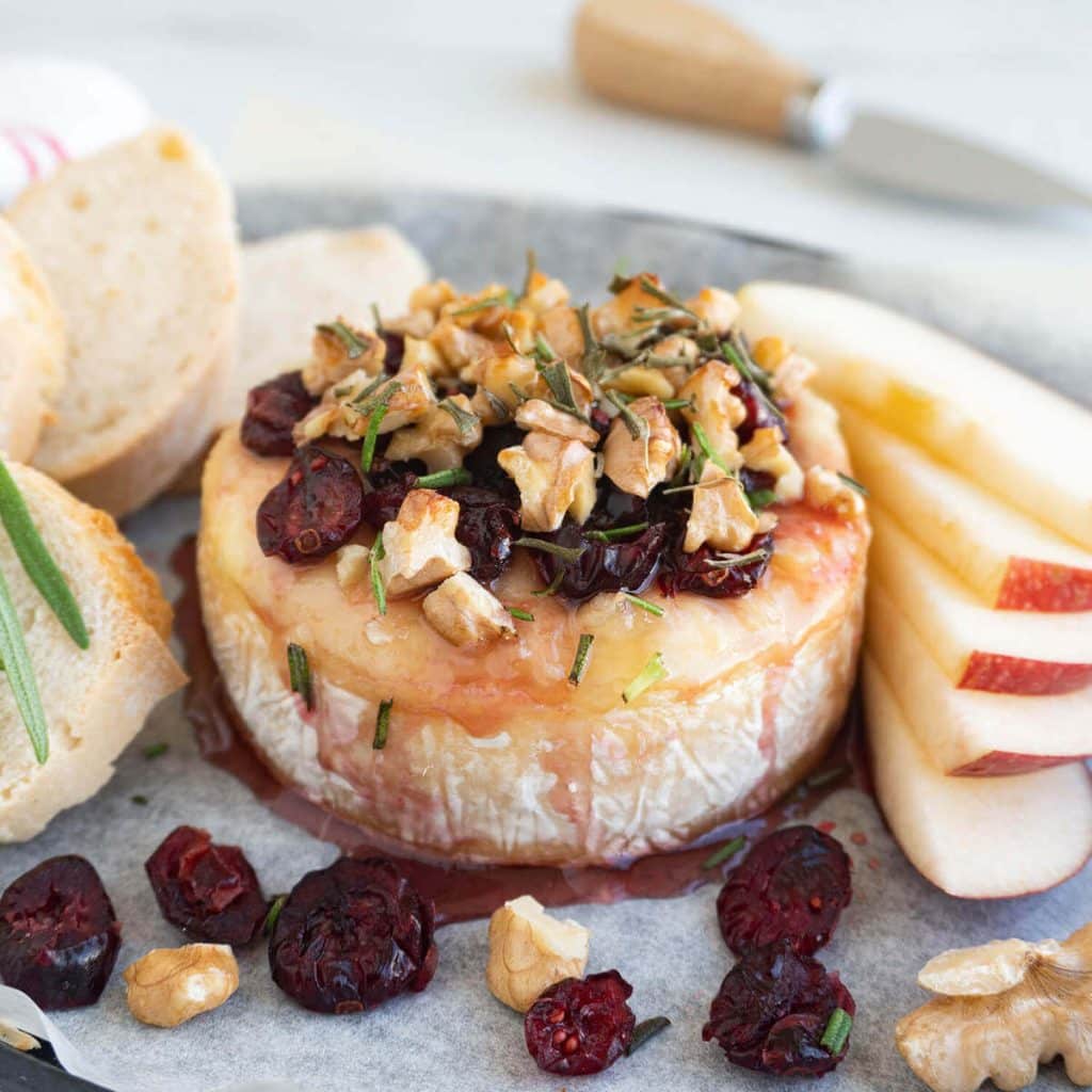 Closeup of baked brie, topped with cranberries, honey, walnuts, and rosemary surrounded by slices of red apples and toasted bread.