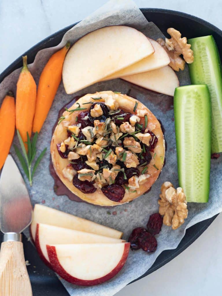 Cranberry baked brie surrounded by apple slices, sliced cucumber, and baby carrots.