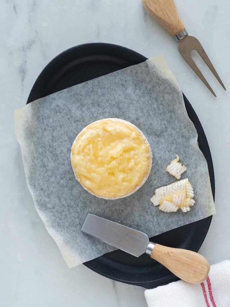 A wheel of brie is on a parchment paper sheet in a baking dish, with the top of the rind shaved off.