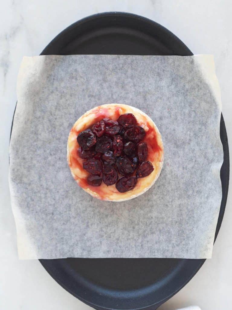 A wheel of brie is on a parchment paper sheet in a baking dish, topped with cranberry sauce, and dried cranberries.