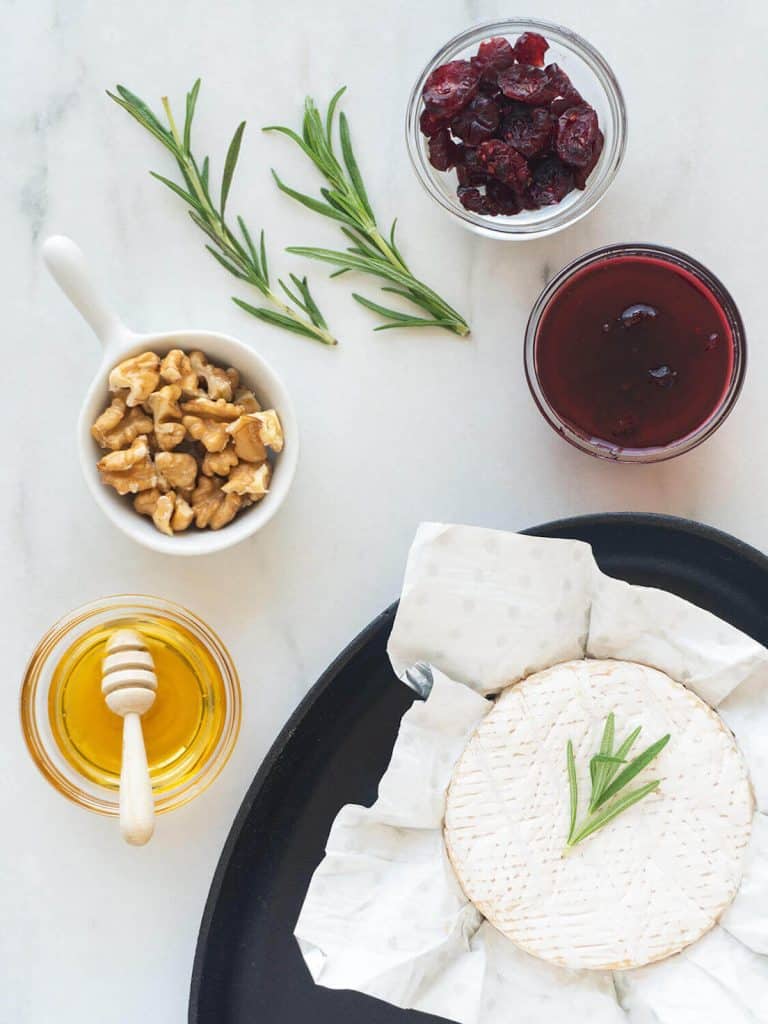 Glass bowls of cranberry sauce, dried cranberries, and honey, along with a white dish of nuts, a brie wheel, and rosemary springs.